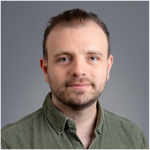 A man with light skin and short brown hair, wearing a green button-up shirt, is facing the camera and smiling softly against a plain grey background.