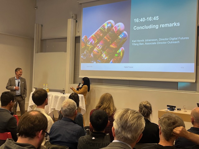 A man stands by a table addressing an audience in a conference room. A woman sits nearby. A presentation slide on the screen reads Concluding remarks with speaker names and a colourful hand graphic.