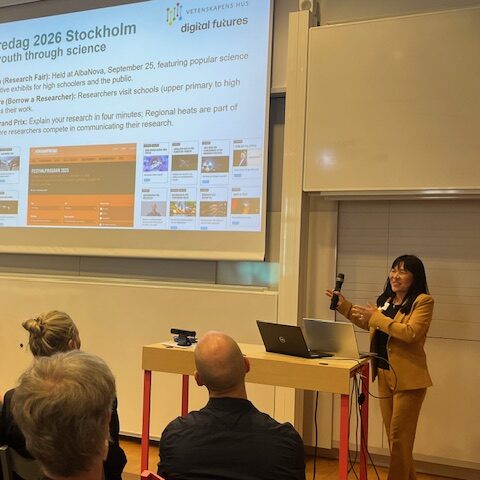 A woman in a yellow blazer presents with a microphone in front of an audience. Behind her, a screen displays a slide titled ForskarFredag 2026 Stockholm with event details and website images.