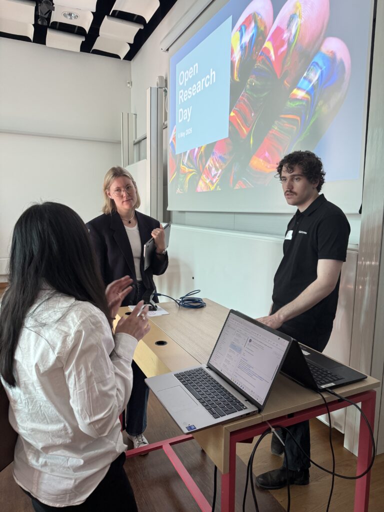 Three people stand and talk near a lectern in a classroom with a large screen behind them displaying Open Research Day and a colourful abstract image. One person uses a laptop on the lectern.