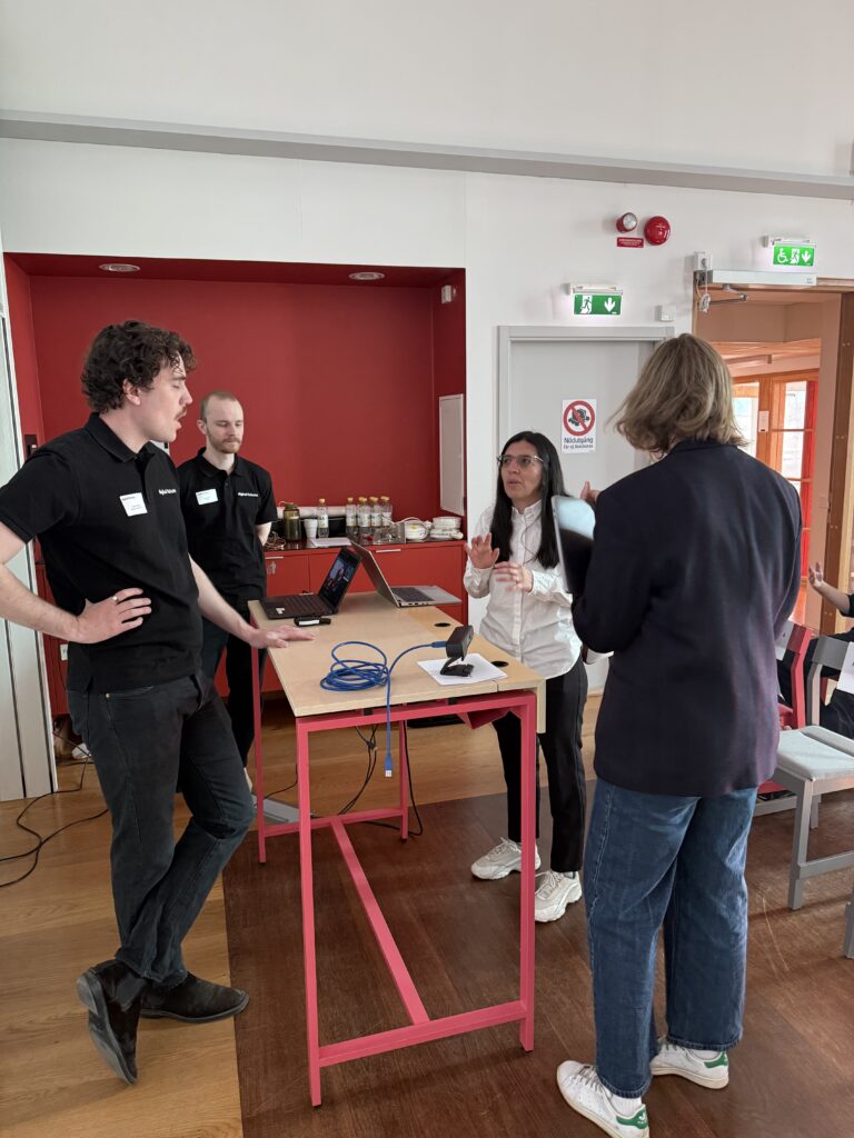Four people are having a discussion around a high table with laptops and cables in a brightly lit room. There are refreshments on a counter in the background and emergency exit signs above the doors.