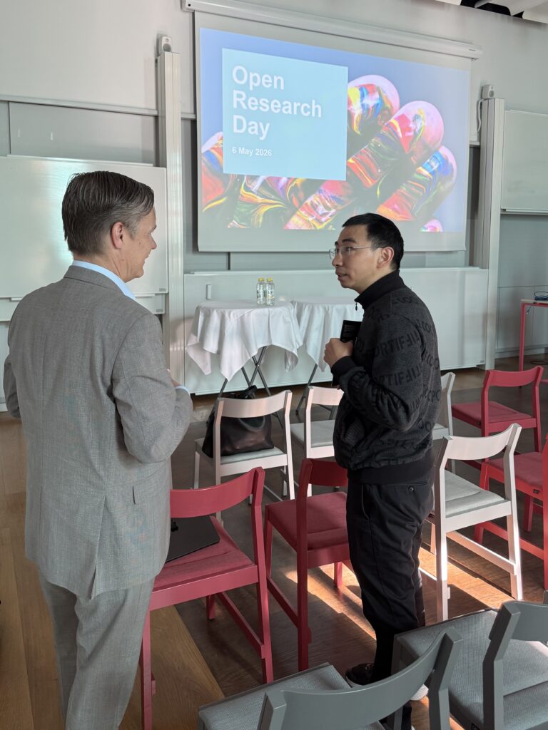 Two men stand and talk in a conference room with empty chairs. Behind them is a screen displaying Open Research Day, 6 May 2026 with colourful light bulbs in the background.