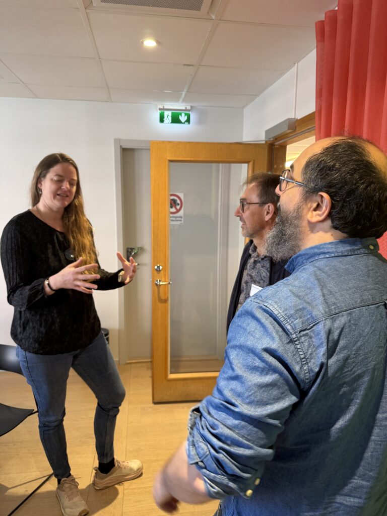 Three people stand and talk in an office. A woman gestures with her hands whilst two men listen. There is a glass door and a red curtain in the background, and the atmosphere appears casual and friendly.