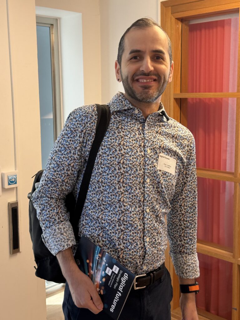 A smiling man with short hair and a beard stands indoors, wearing a patterned shirt and carrying a rucksack. He holds a magazine titled digital futures and has a name badge on his chest. A red curtain and wooden door are in the background.