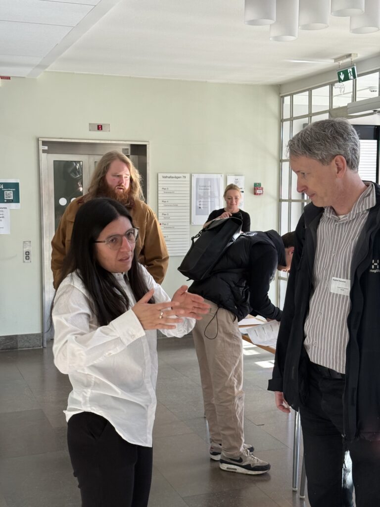A woman in a white shirt gestures while talking to a man in a striped jacket indoors. Two other people are in the background, one writing on a table and another standing nearby with a rucksack.