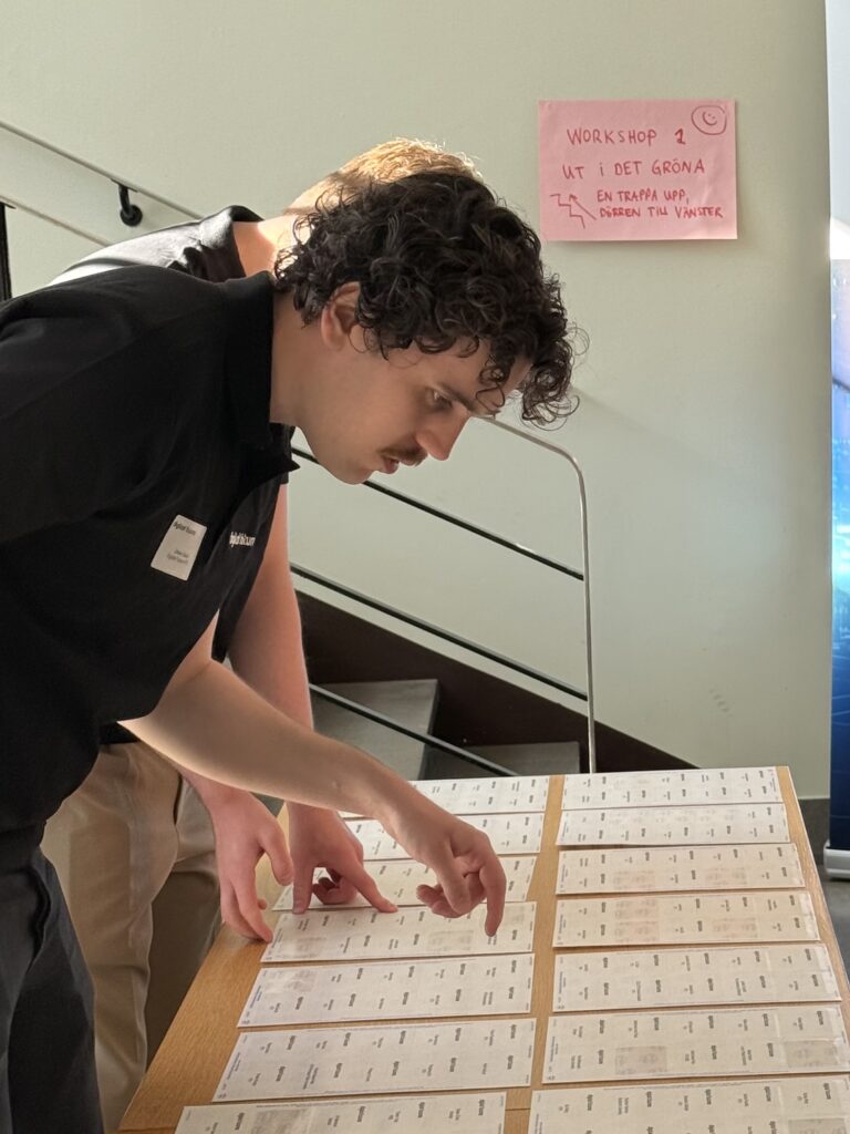 A man with curly hair and a moustache examines name tags laid out on a wooden table. Another person stands beside him. A pink workshop sign is posted on the wall in the background.