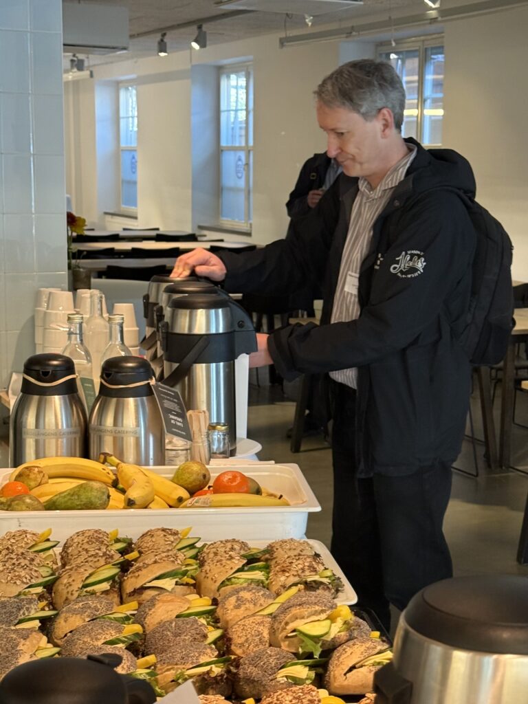 A man in a black jacket pours coffee from a large thermal carafe in a brightly lit room. In the foreground, trays with assorted sandwiches, fruit, and more coffee carafes are on the table.