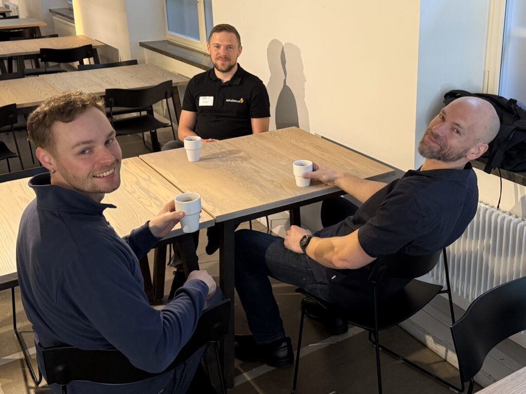 Three men sit around a wooden table in a bright room, smiling at the camera while holding white coffee cups. The table and chairs are simple and black, and sunlight streams in through large windows.
