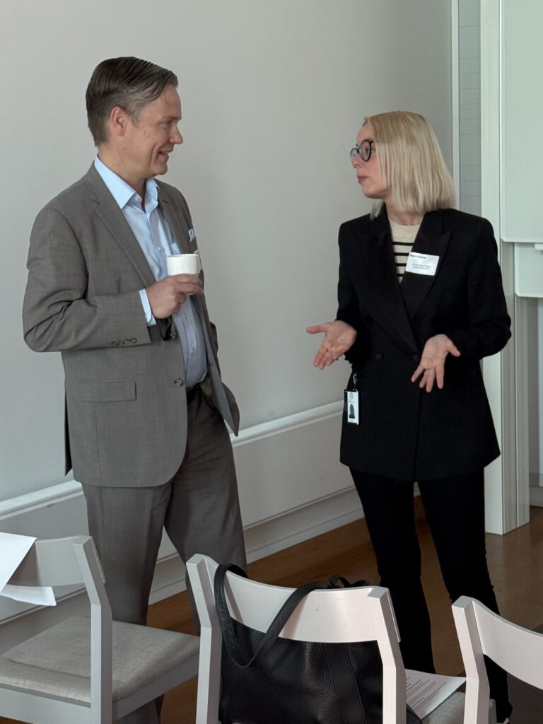 A man in a grey suit and a woman in a black suit and glasses are standing indoors, engaged in conversation, with empty chairs and papers nearby.