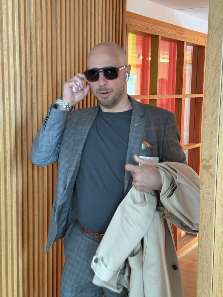 A man in a grey checked suit and sunglasses stands indoors, holding a beige coat over his arm and adjusting his sunglasses, with wooden panelling and colourful glass in the background.