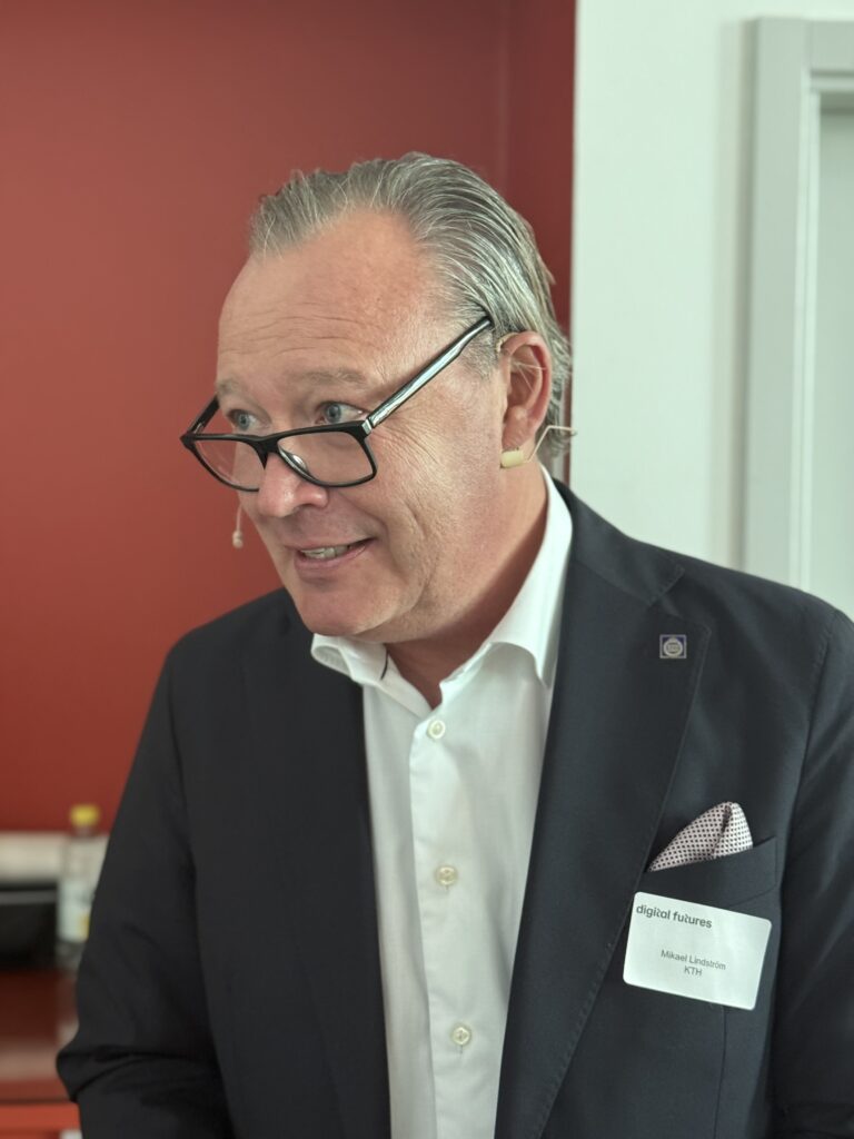 A man in a suit with a microphone headset and glasses speaks, standing indoors by a red wall. He wears a white shirt, pocket square, and a name badge that reads digital futures.