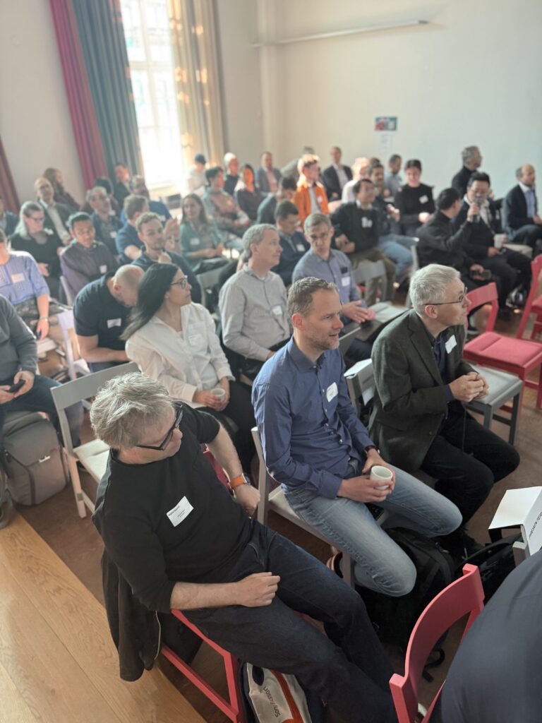 A large group of people seated closely together in a bright room with wooden floors, attending an event or seminar. Most people face forward, listening attentively. Some are holding notebooks or coffee cups.