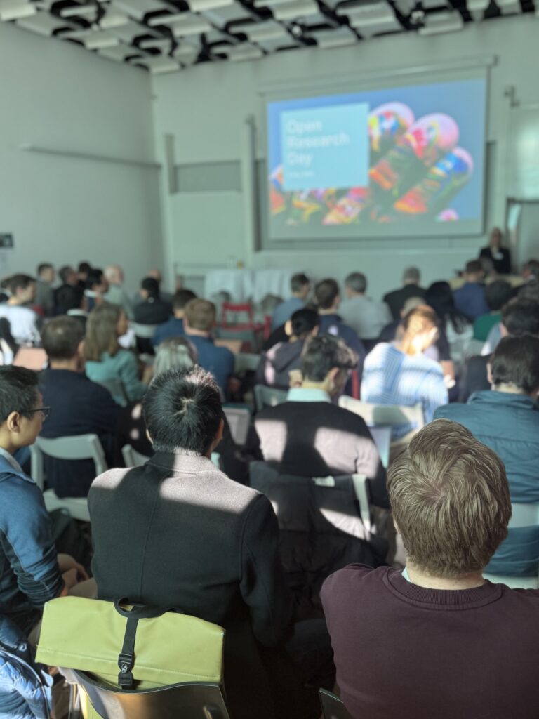 A group of people sit facing a screen in a bright room during a presentation. The screen displays the title “Open Research Day” with colourful graphics in the background.