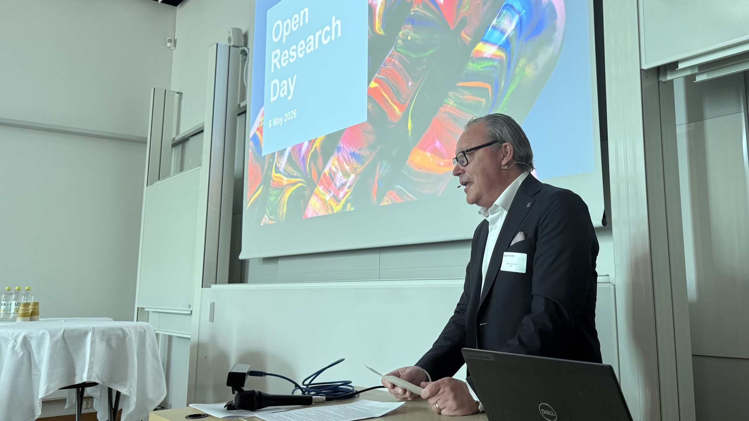 A man in a suit speaks at a lectern in front of a large screen displaying the words Open Research Day and a colourful abstract image, inside a modern conference room.