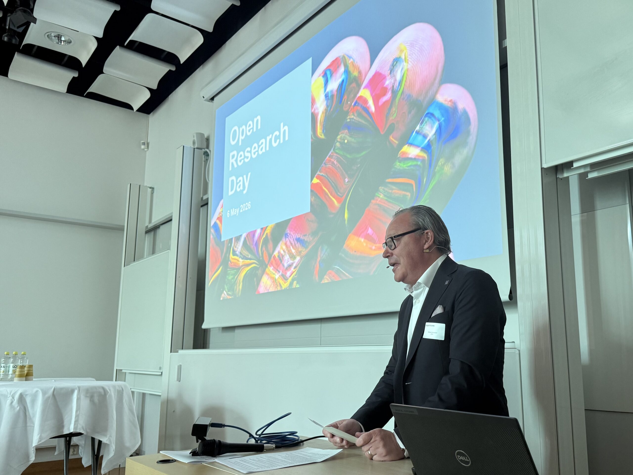 A man in a suit speaks at a lectern in front of a large screen displaying the words Open Research Day and a colourful abstract image, inside a modern conference room.