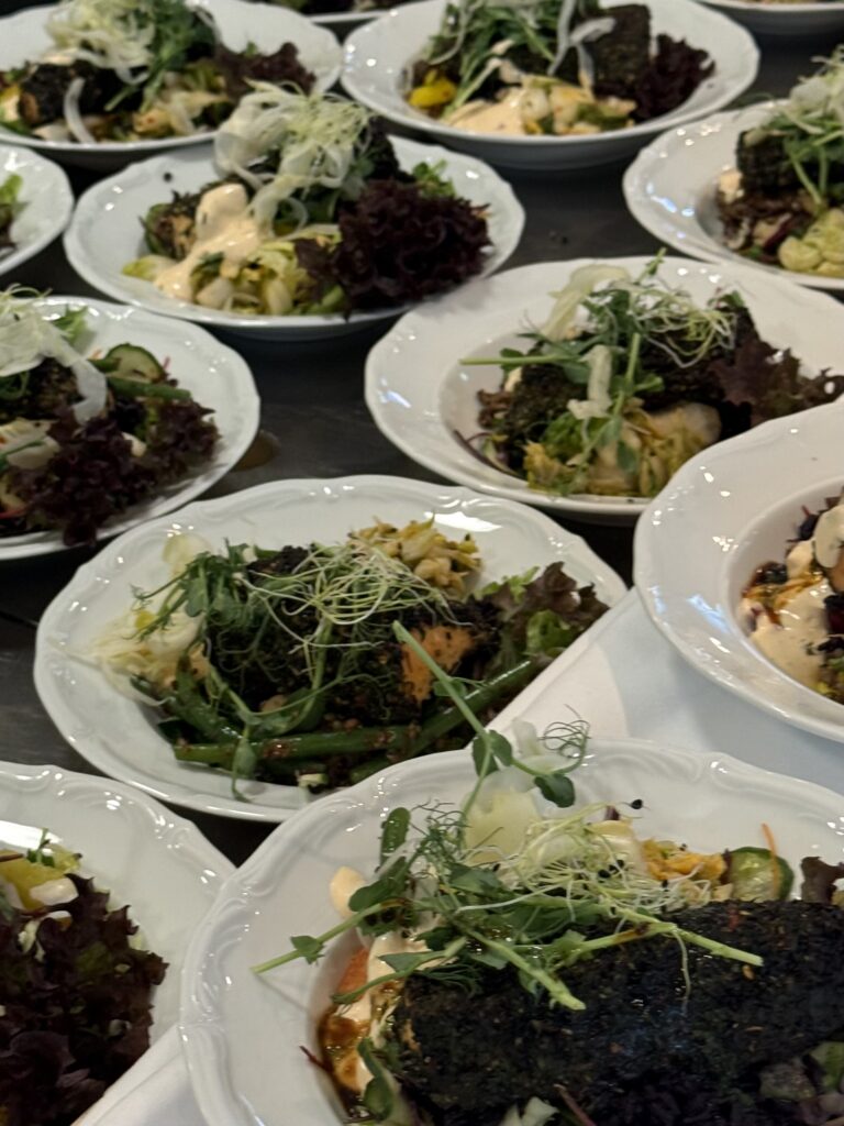 Several white plates arranged on a table, each featuring a gourmet dish with greens, sprouts, leafy lettuce, grilled vegetables, and a piece of herb-crusted protein topped with microgreens.