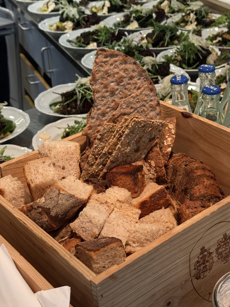 A wooden box filled with assorted breads, including sliced loaves and crispbreads, sits on a table with plates of salad leaves and bottles of water in the background.