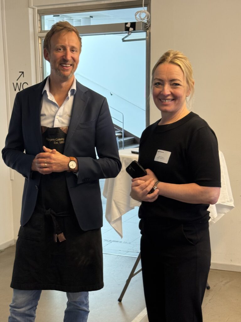 Two people stand indoors, smiling at the camera. The man wears a blue blazer, light shirt, and dark apron, while the woman wears a black outfit and holds a mobile. A table with a white cloth is in the background.