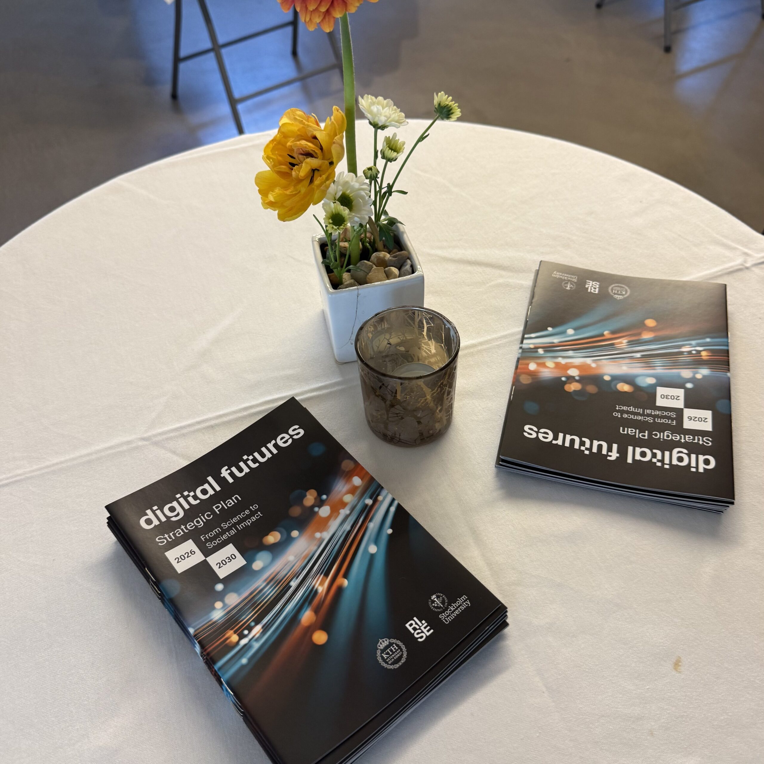 Two digital futures booklets, a glass candle holder, and a small vase with colourful flowers are arranged on a round table covered with a white tablecloth. Empty tables are visible in the background.