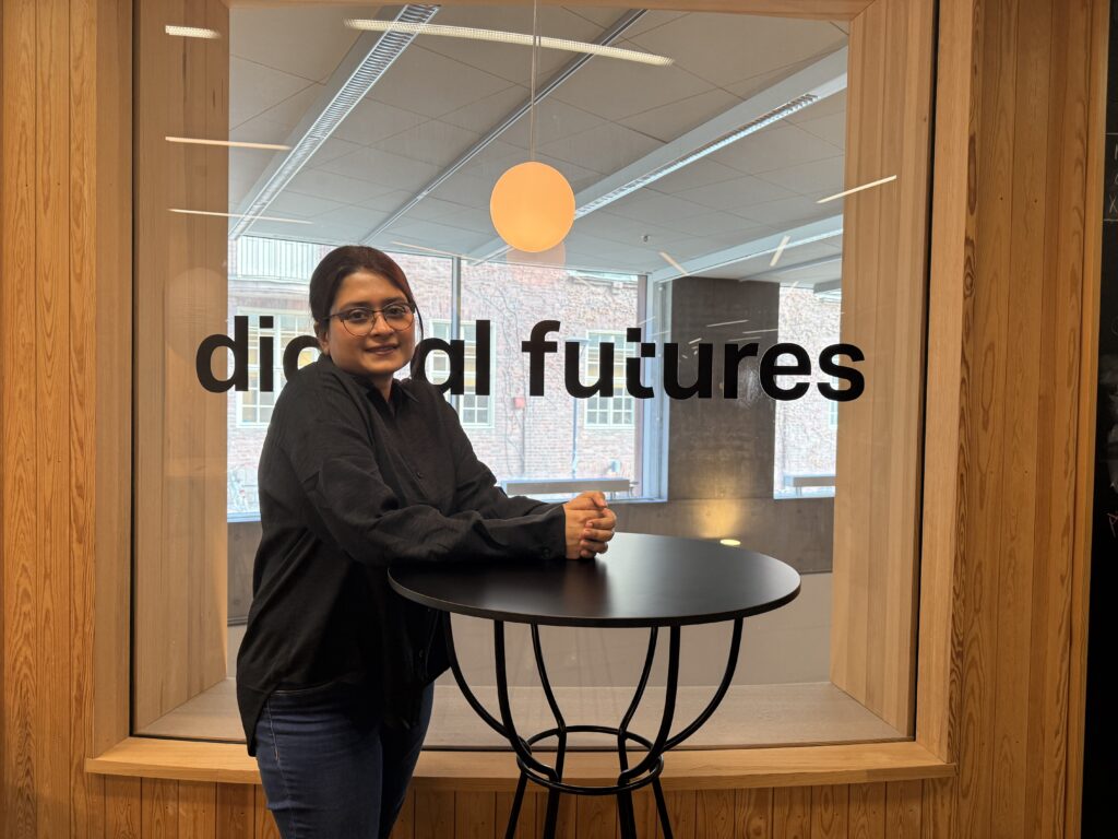 A person with glasses and dark hair stands by a round black table in front of a window with the words digital futures on it, inside a modern building with wooden and glass elements.