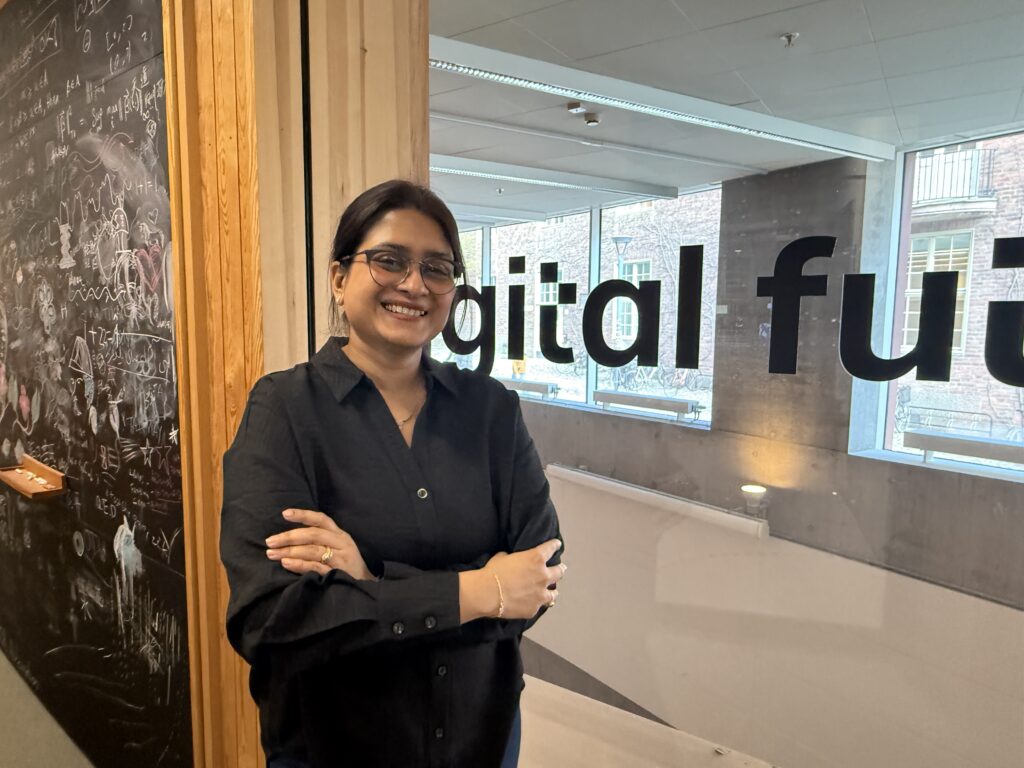 A woman with glasses and a black shirt stands smiling with arms crossed in front of a glass wall displaying the words digital fut. A chalkboard covered in drawings and writing is visible to the left.