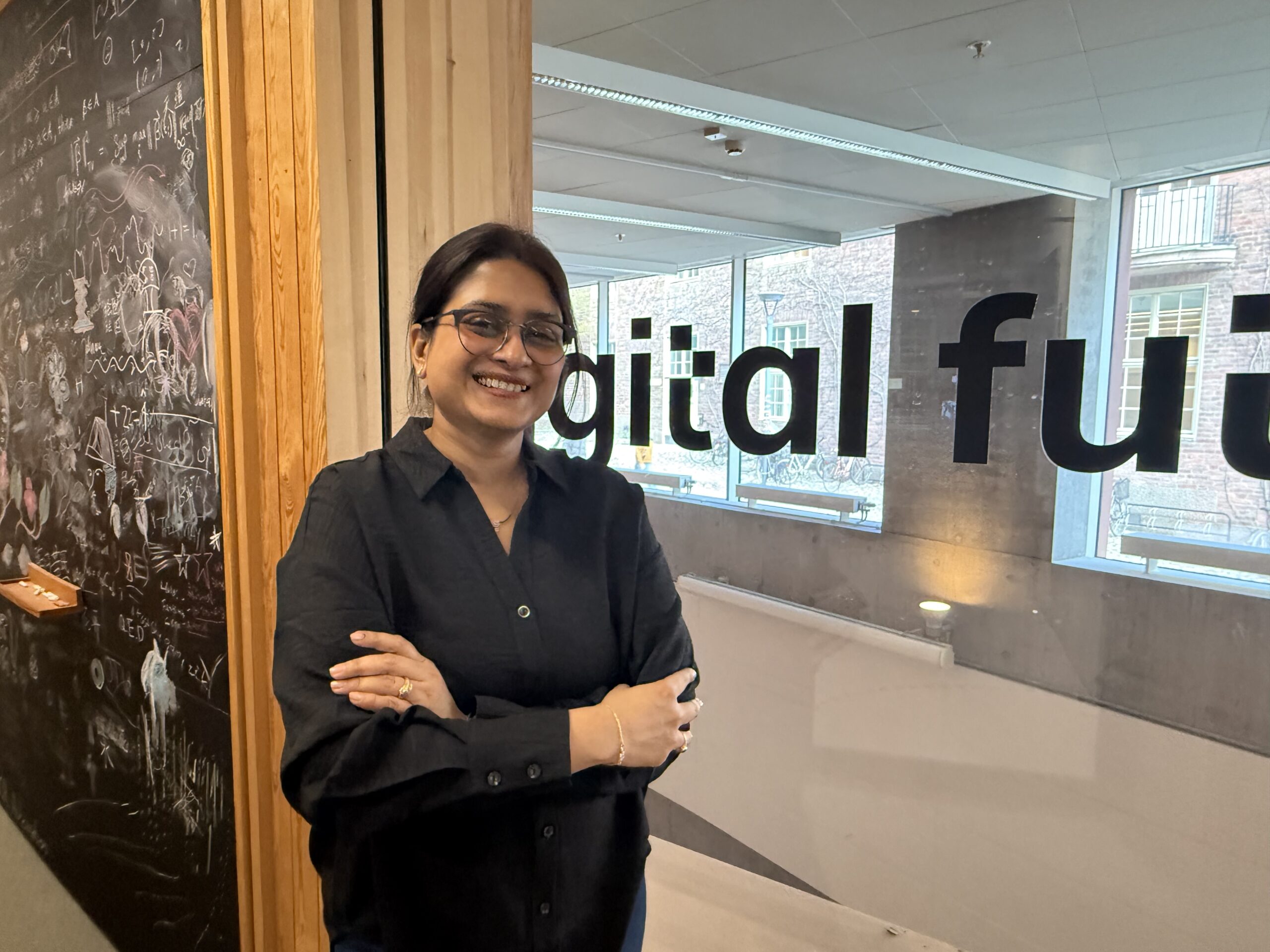 A woman with glasses and a black shirt stands smiling with arms crossed in front of a glass wall displaying the words digital fut. A chalkboard covered in drawings and writing is visible to the left.