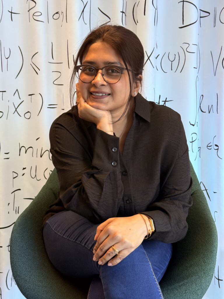 A woman with glasses and a dark shirt sits on a green chair, smiling with her hand resting on her chin. Mathematical formulae are written on the white background behind her.