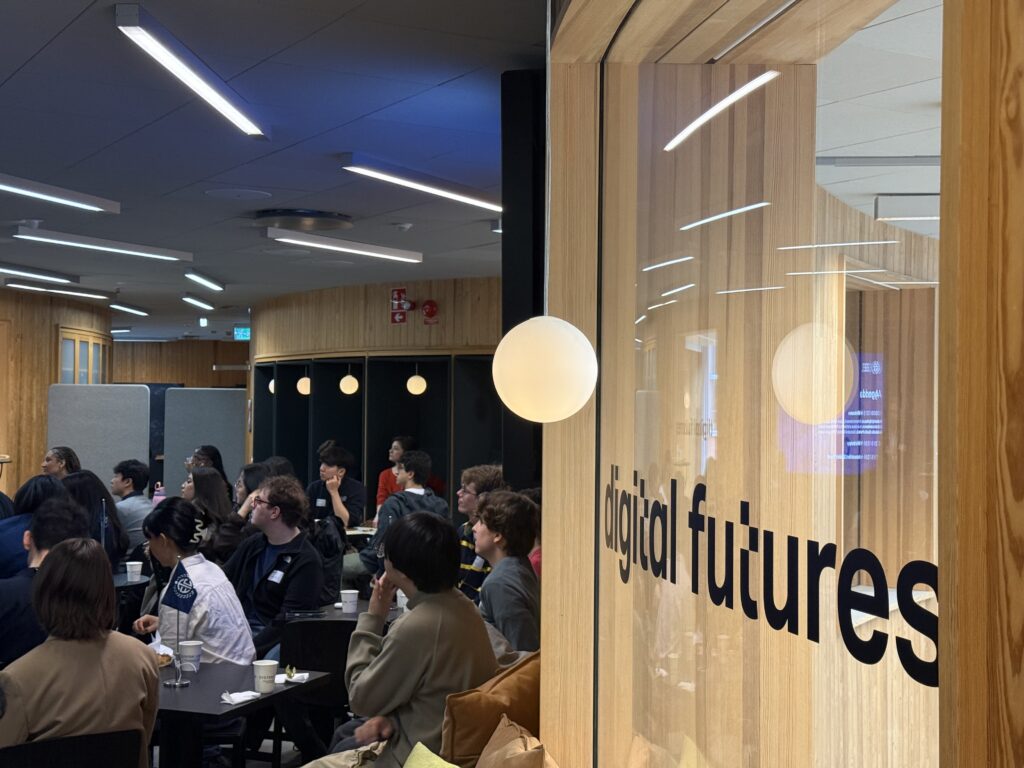 A group of people sit in a modern room with wooden walls and large glass panels. The words digital futures are written on the glass, and attendees face a screen, suggesting a presentation or event.