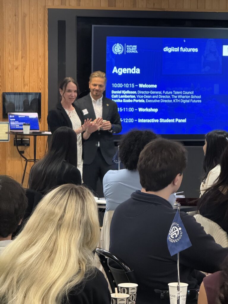 A woman and a man stand at the front of a room, addressing a seated audience. Behind them is a large screen displaying an event agenda for a workshop, and a blue flag with a white logo is visible in the foreground.