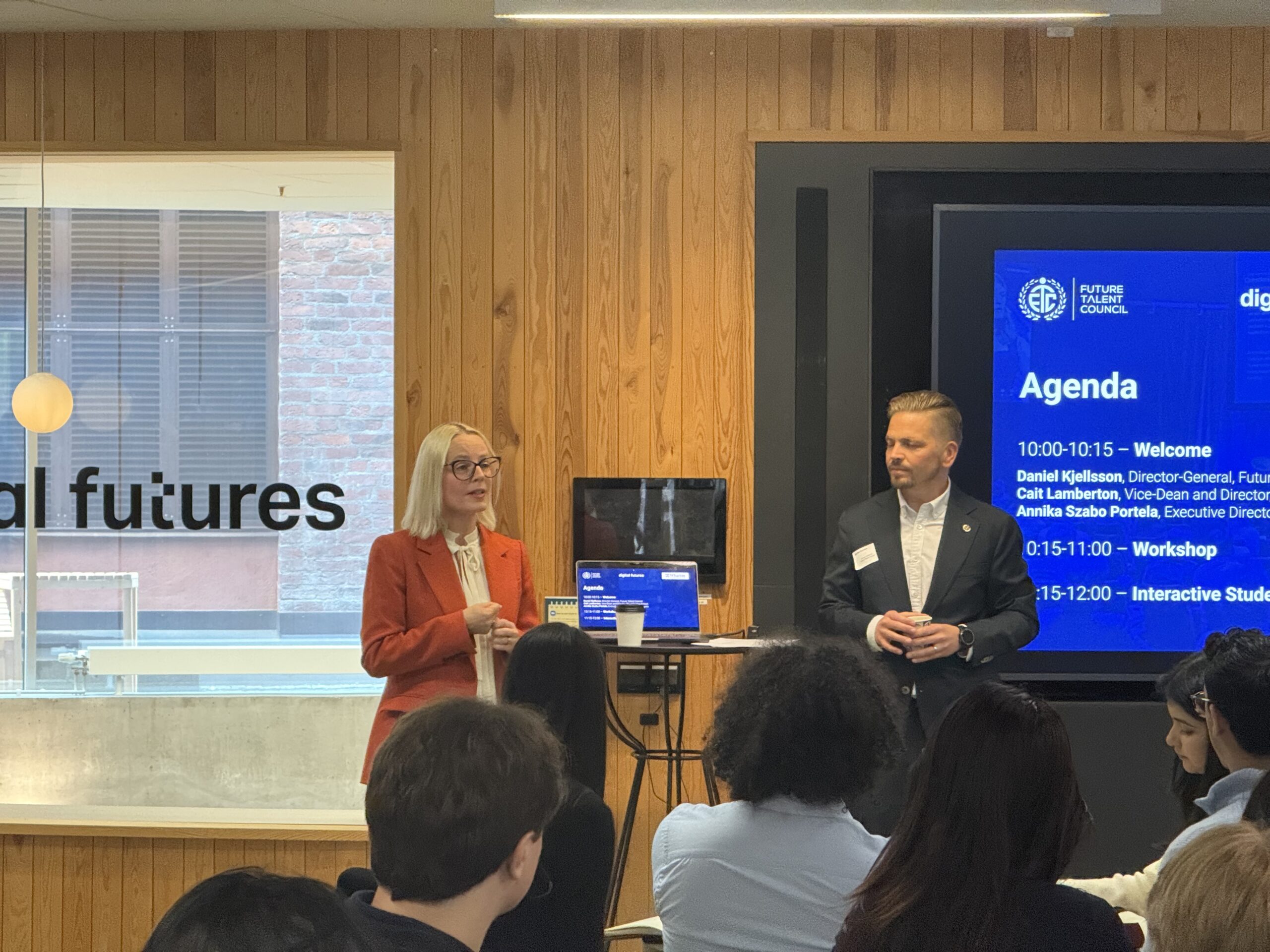 Two presenters stand at the front of a room speaking to an audience. Behind them, a large screen displays an agenda with event details. The room has wood panelling and large windows.