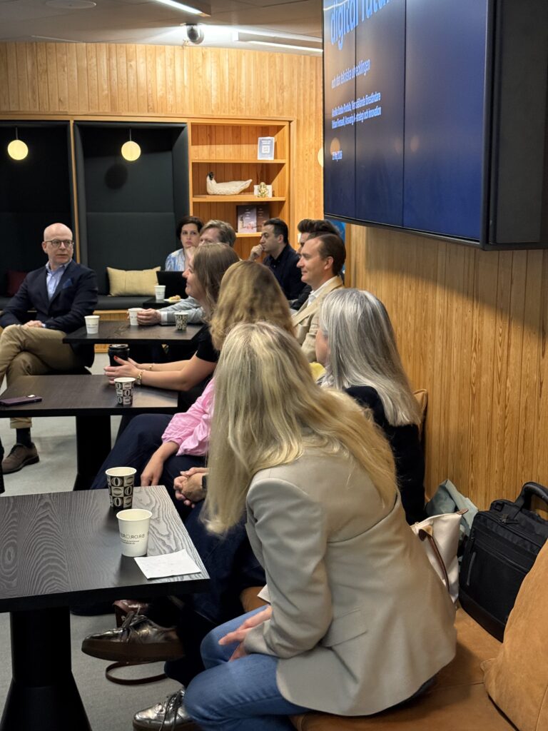 A group of people sit around tables in a modern meeting room, listening to a man in glasses who appears to be leading a discussion. A large screen is visible on the wall, displaying text.