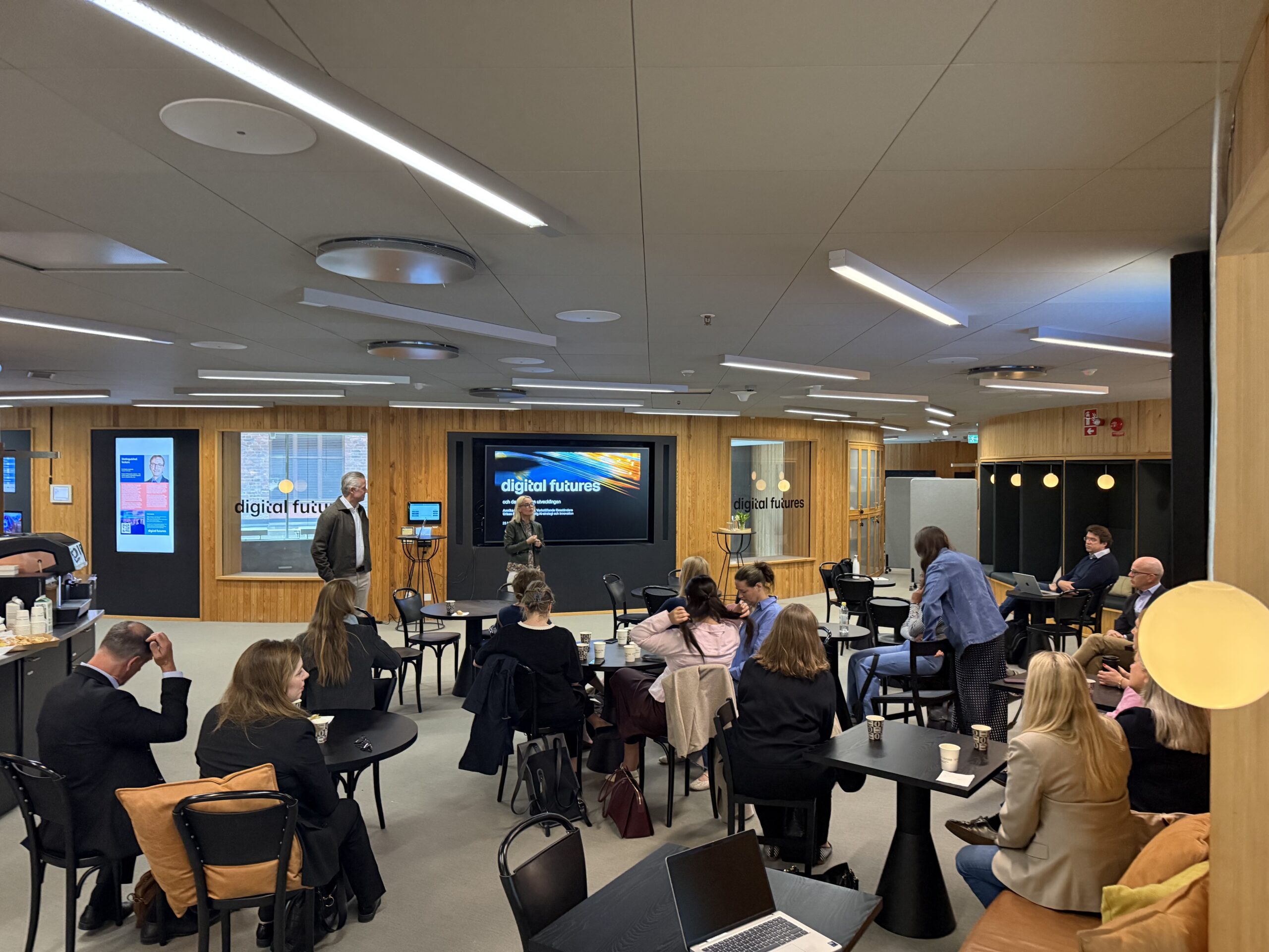 People are seated at tables in a modern conference room, attentively listening to a presentation titled digital futures displayed on a large screen at the front. The atmosphere is professional and collaborative.