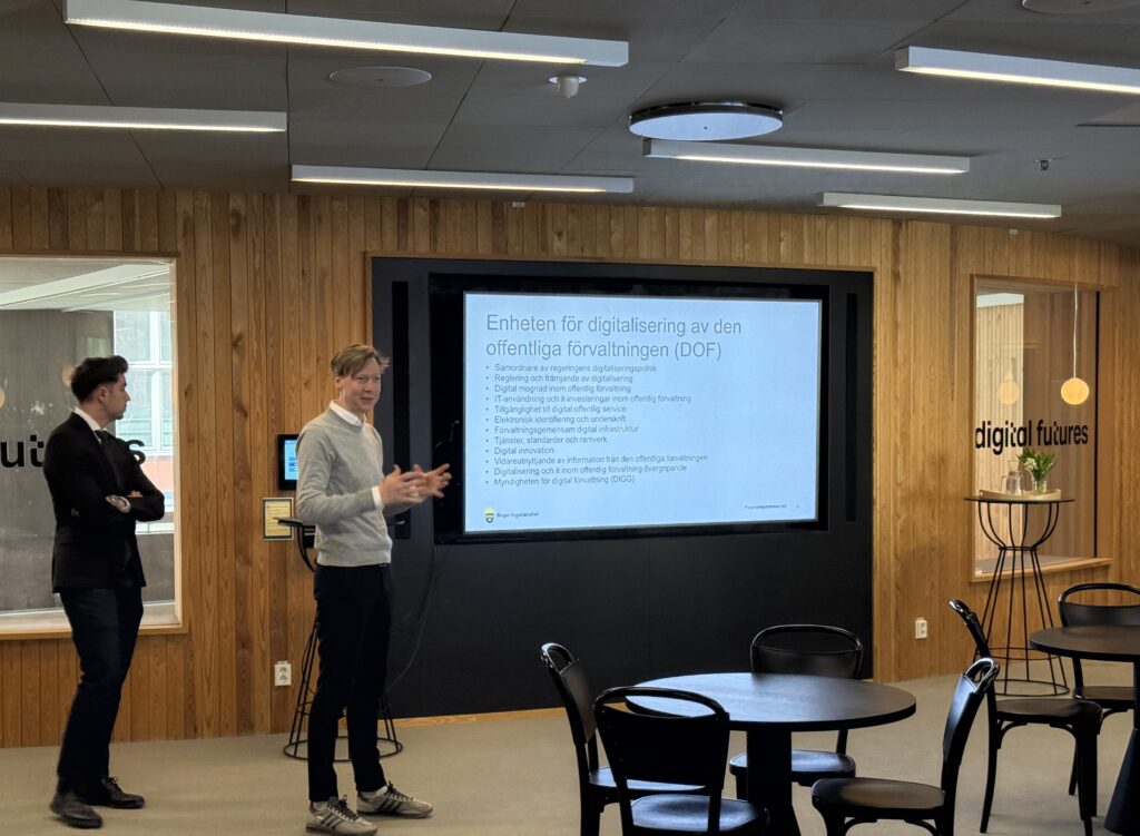 Two people stand in front of a screen giving a presentation in a modern office space. The screen displays Swedish text about digitalisation in public administration. Empty chairs and tables are visible in the foreground.