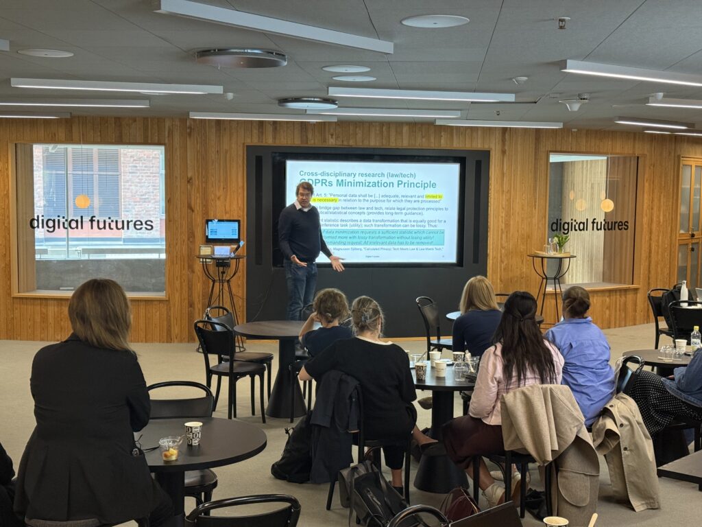 A man stands and presents in front of a screen displaying a slide titled PRS Minimisation Principle to an audience seated at tables in a modern room with digital futures signs on the walls.