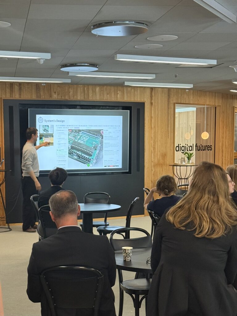 A person presents a slide on system design to an audience in a modern office space with wooden walls and a “digital futures” sign near glass doors. People are seated and facing the presenter.