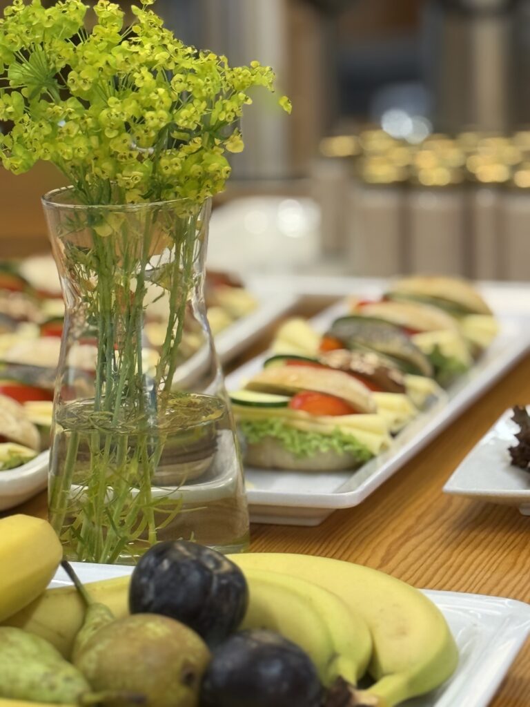 A clear glass vase with yellow flowers sits on a wooden table next to platters of assorted sandwiches with lettuce, cheese, and tomato, and a plate of fresh fruit including pears, plums, and apples.