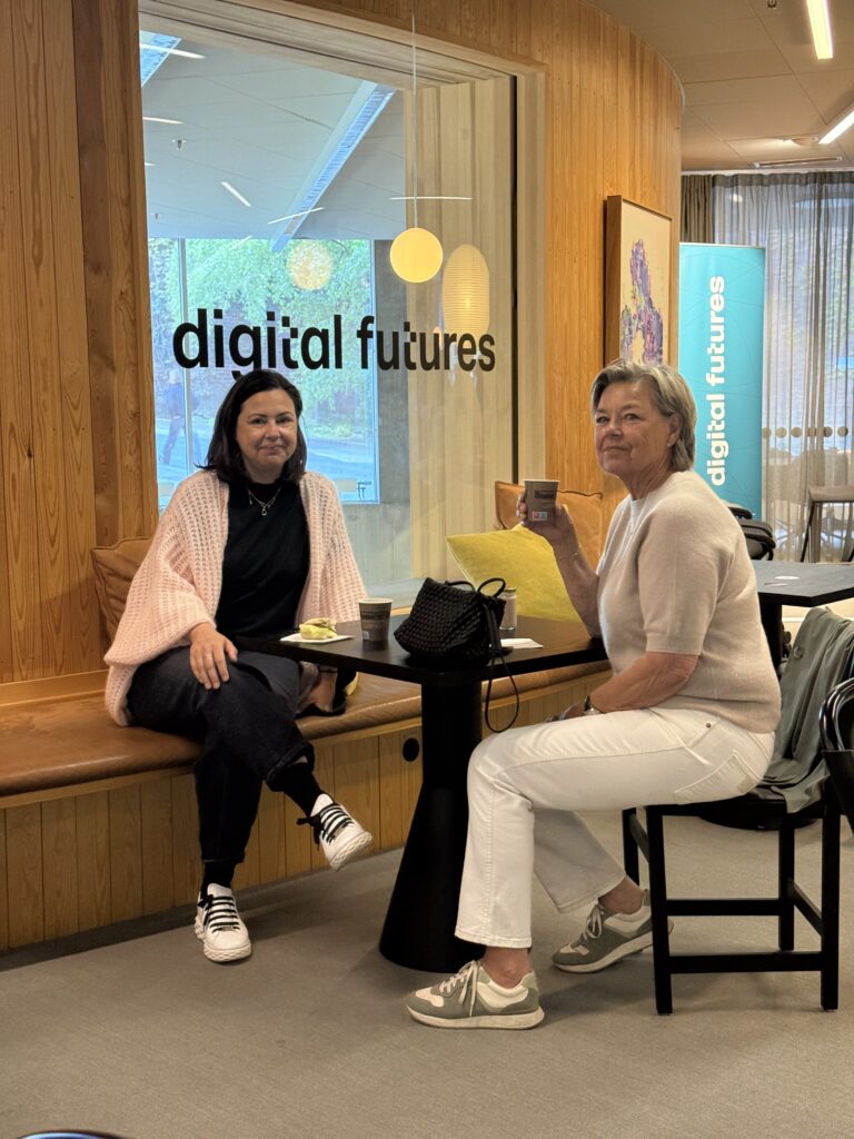 Two women sit at a small black table, smiling and drinking coffee, in a modern space with wooden walls. Behind them, a glass wall displays the text “digital futures”.