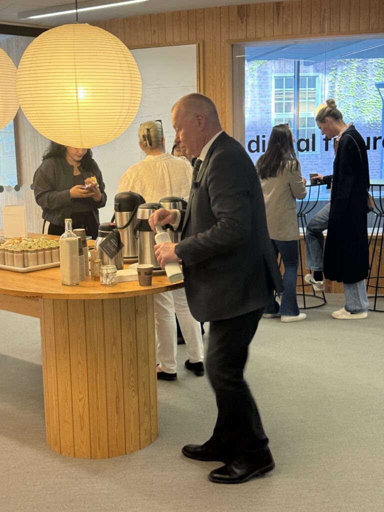 A man in a suit pours coffee at a round wooden table with snacks and drinks, while four other people stand and talk near a window in a modern, bright office space.