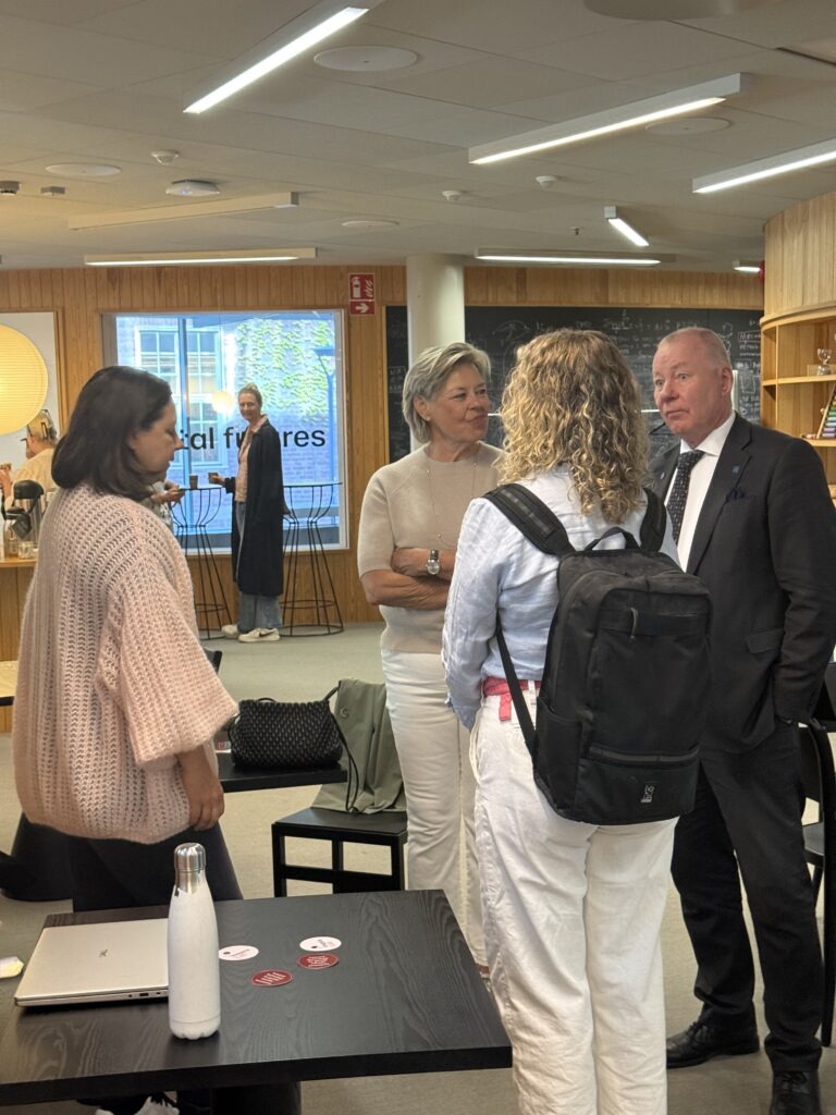 Four people stand in a modern, well-lit office space, engaged in conversation. A woman in the background is seated by a window with a “social futures” sign visible outside. Laptops and water bottles are on a table nearby.