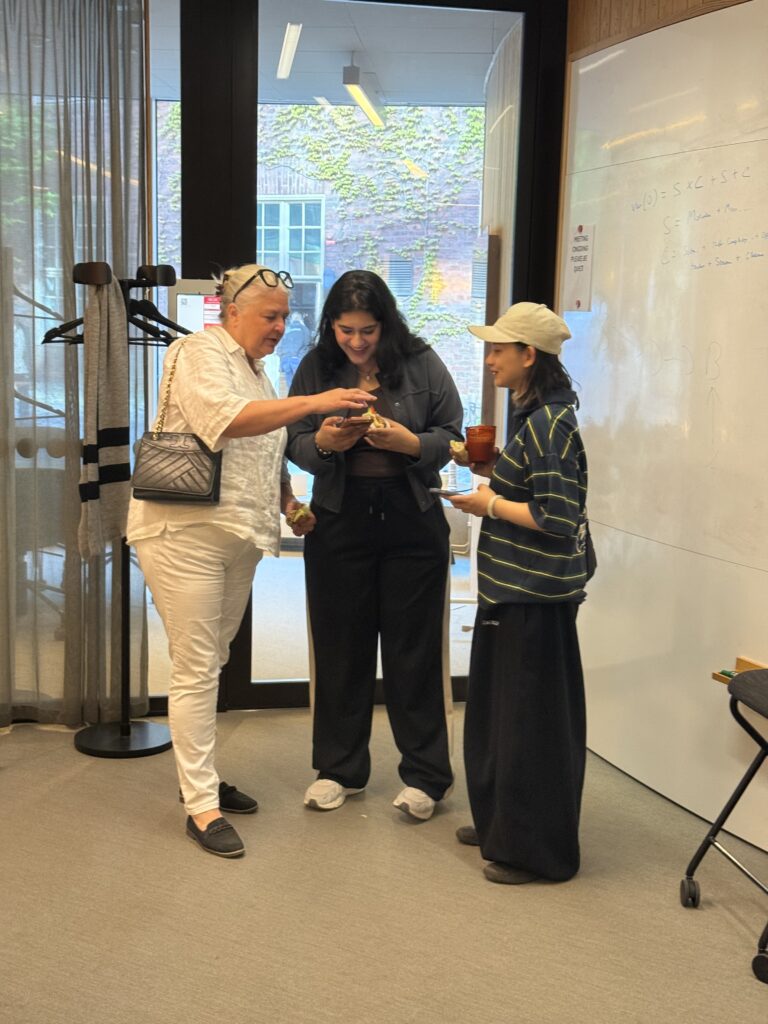 Three people stand together indoors, smiling and looking at a mobile. They appear relaxed and are holding drinks. Behind them is a whiteboard, coat rack, and large window showing greenery outside.