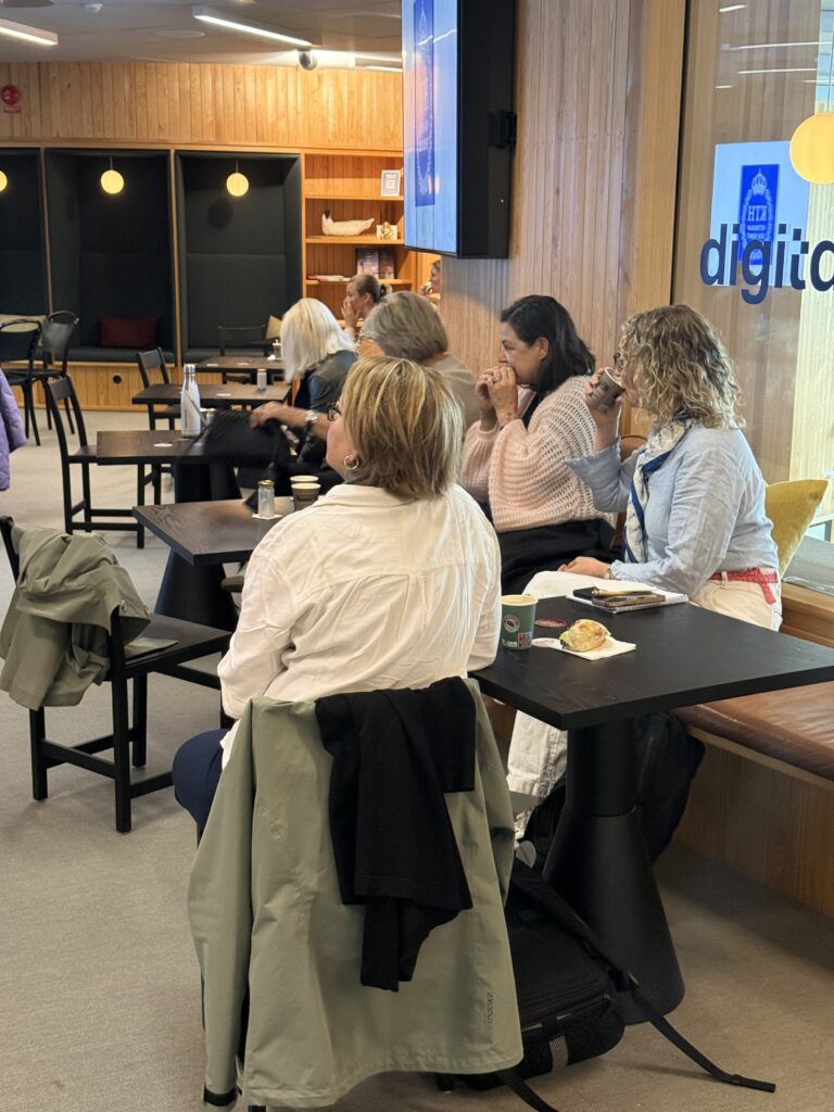 A group of people sit at tables in a modern, wooden-panelled room, listening attentively. Some have notebooks and drinks. The atmosphere is casual and focused, with jackets draped over chairs and natural light coming in.