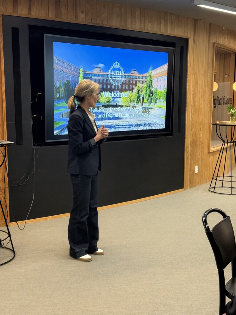 A woman in a blazer and jeans stands and speaks in front of a screen displaying Welcome! and an image of a large building, inside a modern room with wooden walls and high tables.