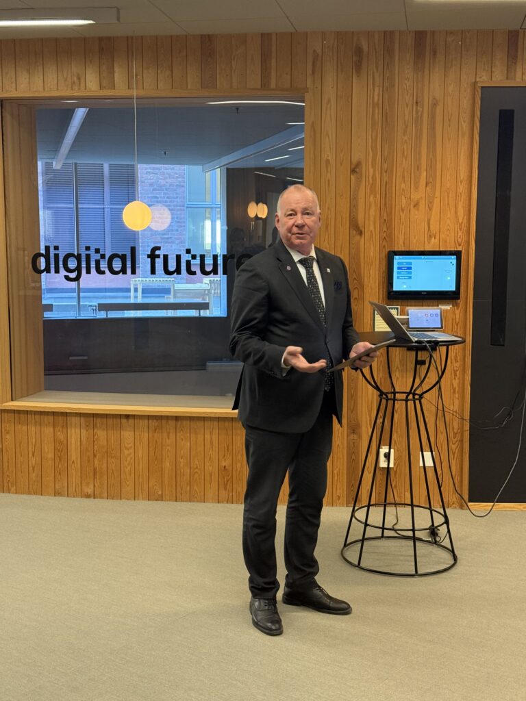 A man in a suit stands next to a high table with a laptop, gesturing with one hand. Behind him, a glass wall displays the words digital future in a modern office setting with wood panelling.