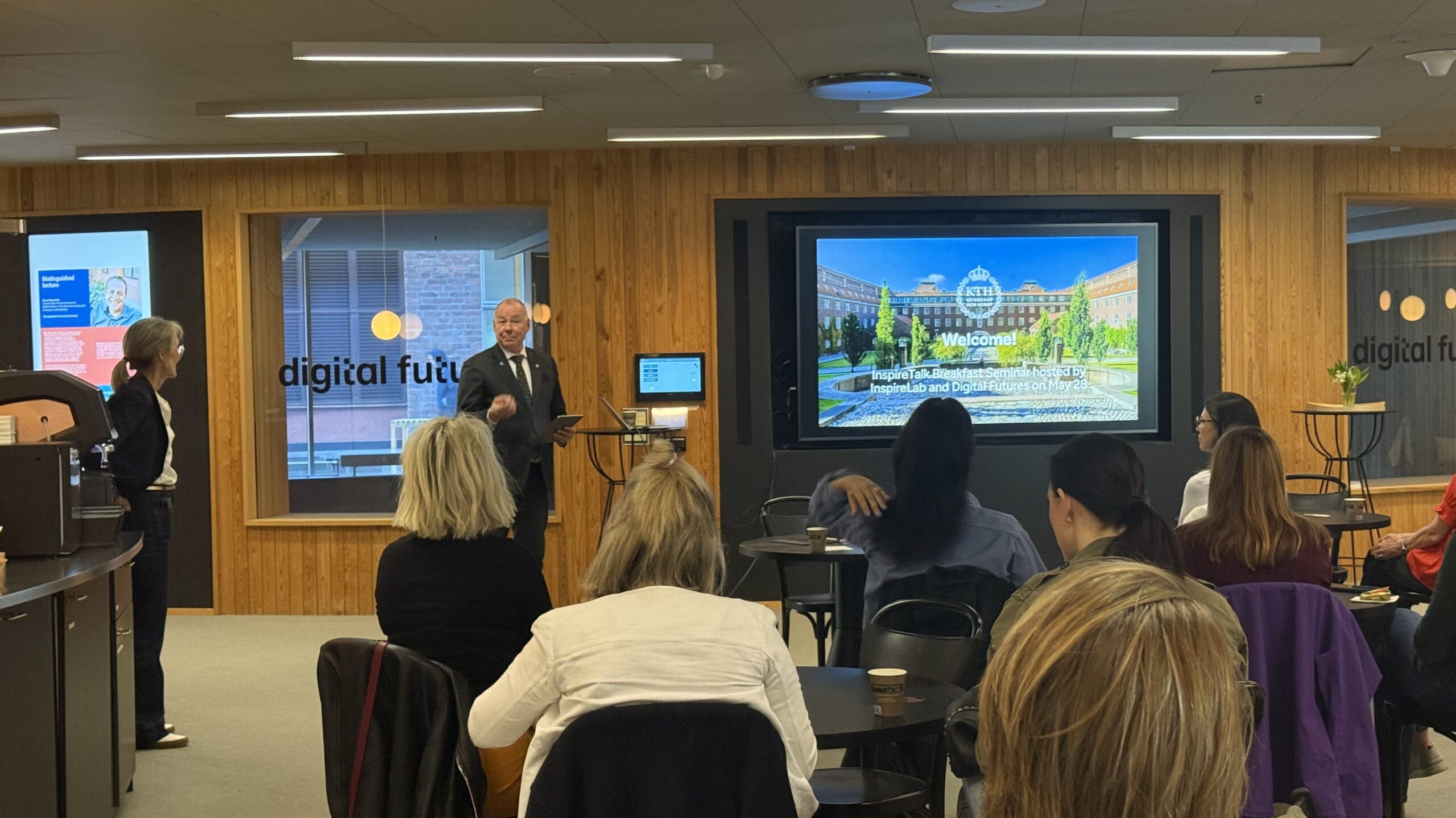 A man in a suit talks to a seated audience in a modern conference room with wood-panelled walls. A large screen behind him displays a welcome slide with a campus image and the words WELCOME.