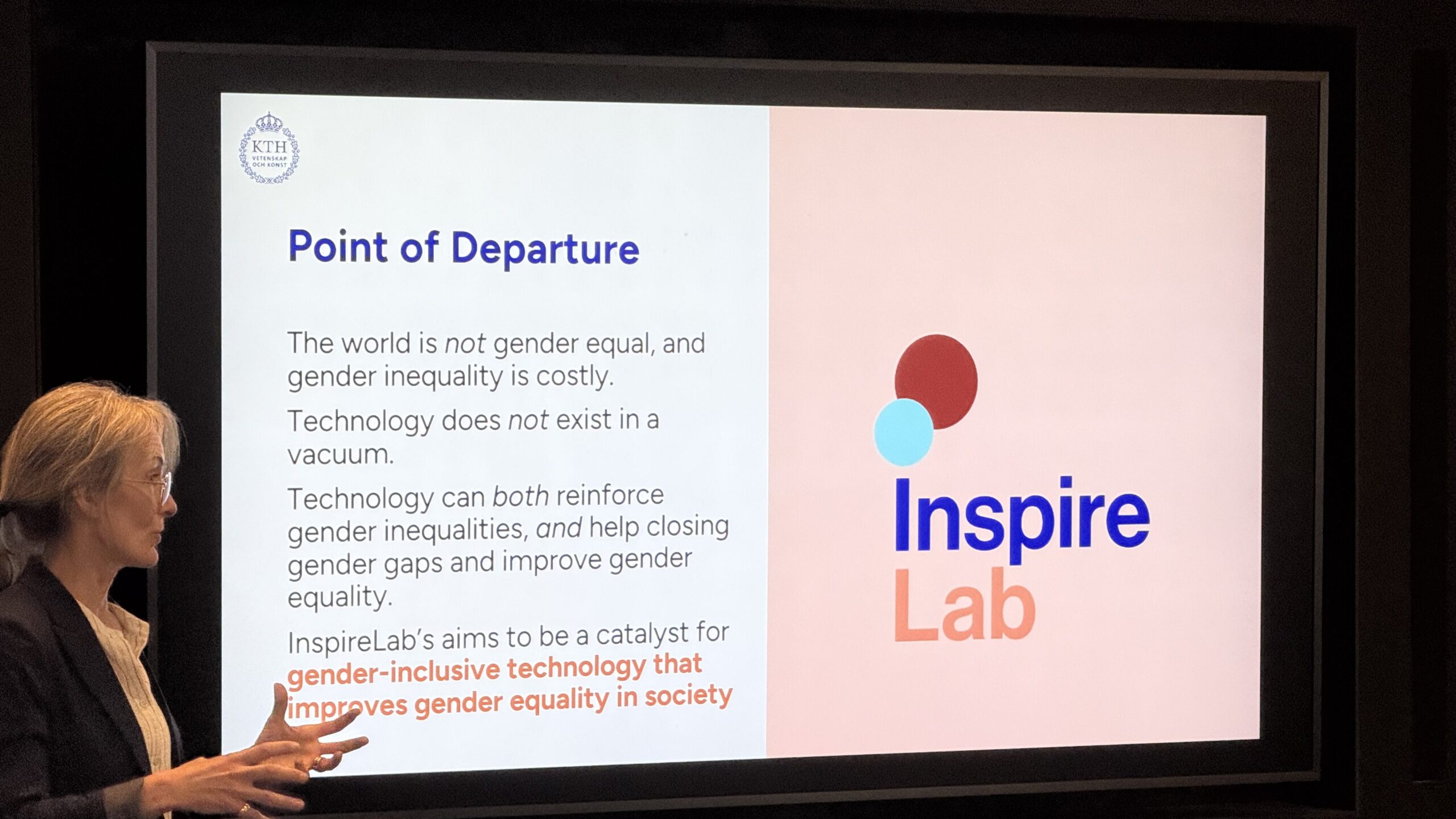 A woman presents a slide titled Point of Departure with text about gender, technology, and equality, next to the Inspire Lab logo on a large screen. Two people are visible in the foreground, listening.