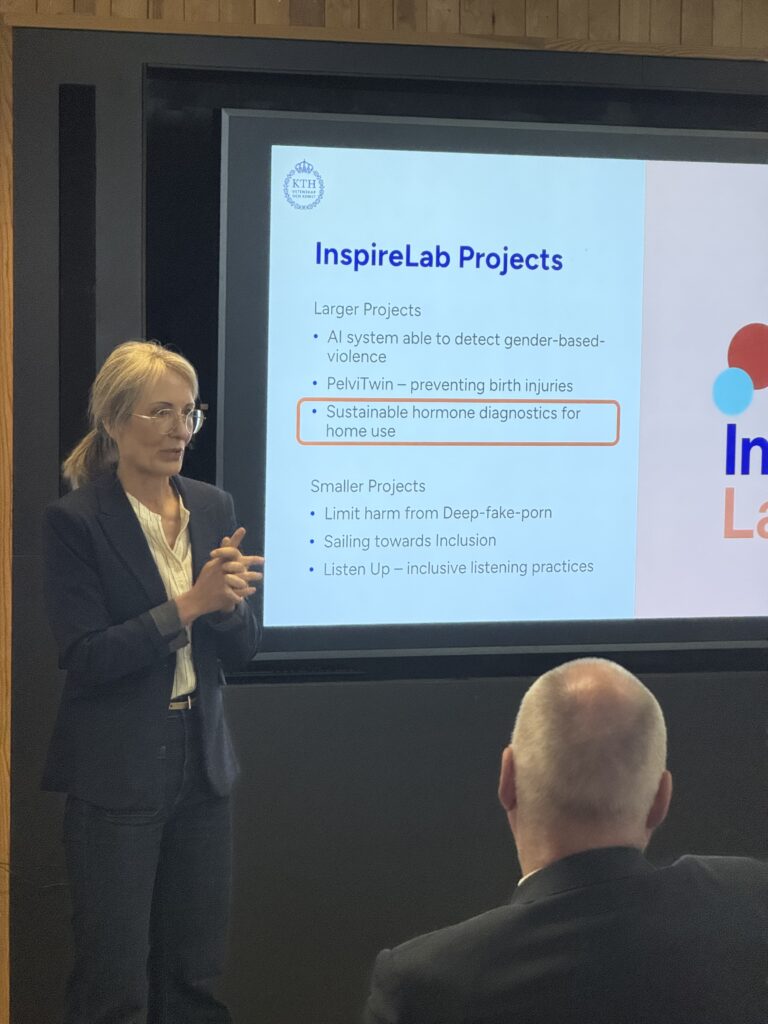A woman in a suit stands and speaks in front of a screen displaying a list of InspireLab Projects to an audience, with project titles including AI for gender-based violence detection and hormone diagnostics.