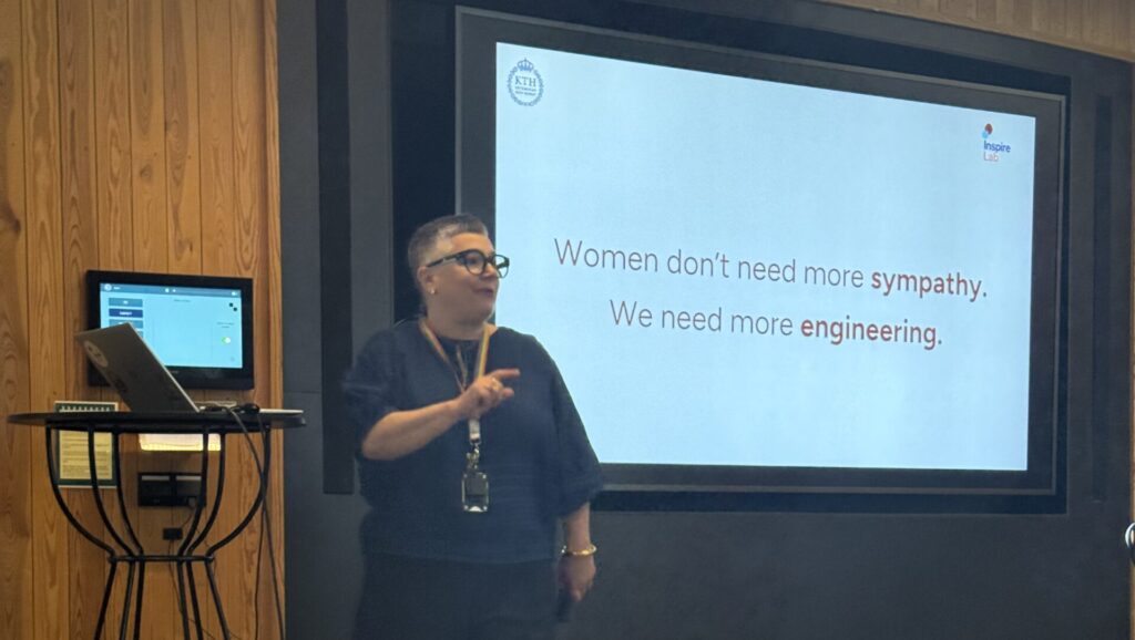 A person stands in front of a screen giving a presentation. The slide reads, Women don’t need more sympathy. We need more engineering. A laptop sits on a small stand nearby. The setting appears to be an indoor meeting room.