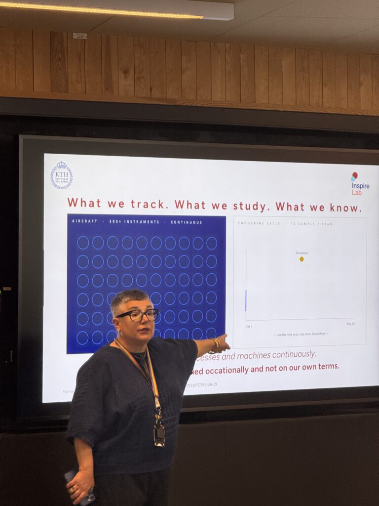 A woman stands in front of a screen giving a presentation. The slide behind her shows a diagram with blue circles on the left and a chart with a yellow dot on the right, along with the title What we track. What we study. What we know.