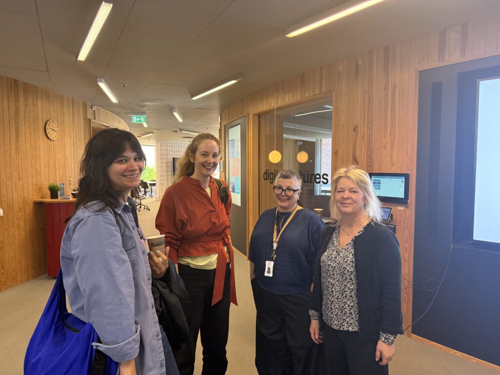 Four women smiling and standing together in a modern office corridor with wooden walls and glass doors. One holds a coffee cup, and two are wearing lanyards. A clock and some plants are visible in the background.
