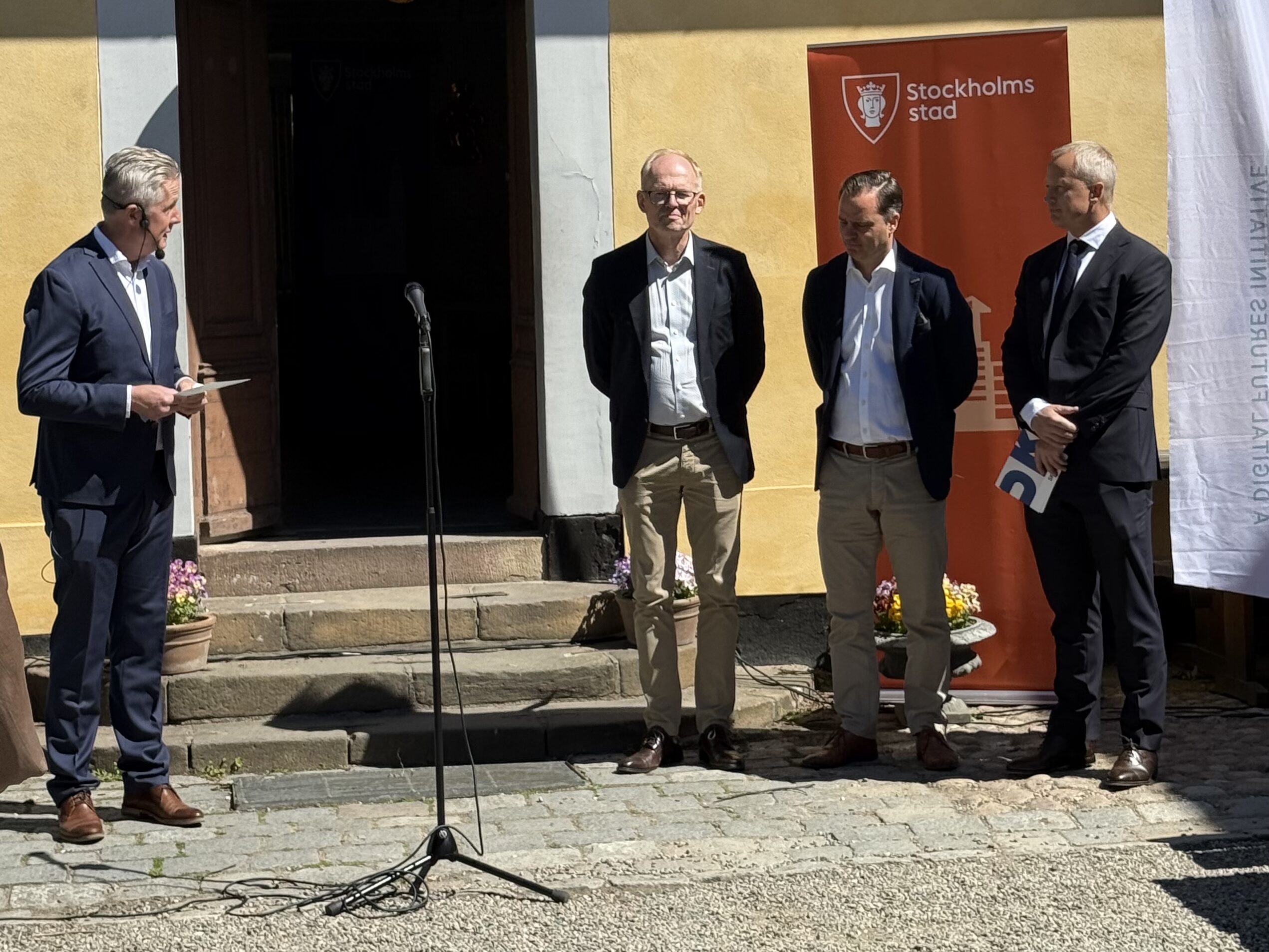 Four men in suits stand outside a yellow building with a tiled roof, one holding a notepad. A microphone stand is placed in front of them, and a banner with Stockholms stad is visible in the background.
